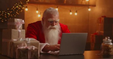 Noel Baba oturma odasında dizüstü bilgisayar kullanıyor. Düşünceli Noel Baba 'nın portresi dekore edilmiş evde modern bir dizüstü bilgisayarla daktilo ediyor.