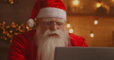 Noel Baba oturma odasında dizüstü bilgisayar kullanıyor. Düşünceli Noel Baba 'nın portresi dekore edilmiş evde modern bir dizüstü bilgisayarla daktilo ediyor.