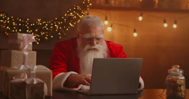 Skutečný Santa Claus pomocí nové technologie pro komunikaci s dětmi, příjem pošty nebo seznam přání. Veselý pracuje na notebooku a usmívá se, zatímco sedí u svého křesla s krbem a vánoční strom — Stock video
