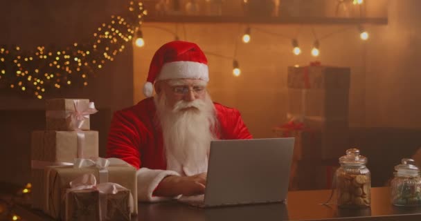 Санта Клаус использует ноутбук в гостиной. Портрет вдумчивого Деда Мороза, печатающего с современным ноутбуком в украшенном доме — стоковое видео