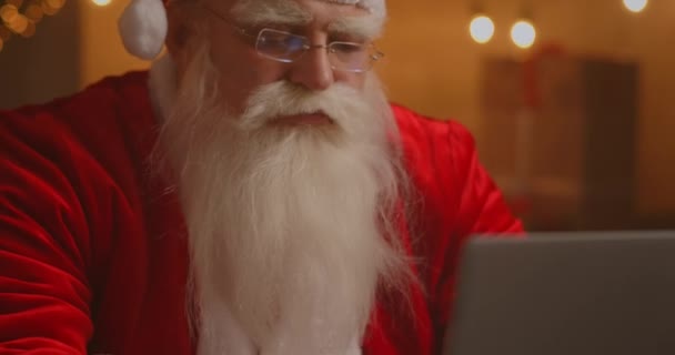 Le vrai Père Noël utilise les nouvelles technologies pour communiquer avec les enfants, recevoir du courrier ou une liste de souhaits. Joyeux travail sur ordinateur portable et souriant tout en étant assis à sa chaise avec cheminée et arbre de Noël — Video