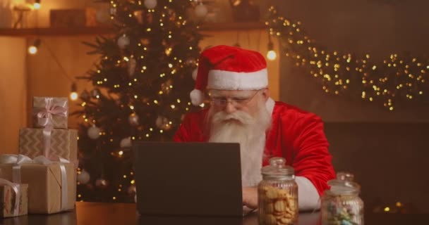 Санта-Клаус держит подарочную коробку набирая клавиатуру ребенок разговаривает с ребенком поздравления на Рождество, с Новым годом в виртуальном онлайн чате на ноутбуке сидя за столом поздно с подарком на Рождество накануне. — стоковое видео