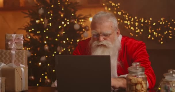 Santa Claus Nyata menggunakan teknologi baru untuk komunikasi dengan anak-anak, menerima surat atau daftar keinginan. Ceria bekerja di laptop dan tersenyum sambil duduk di kursinya dengan perapian dan Pohon Natal — Stok Video