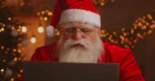 Echte Kerstman met behulp van nieuwe technologie voor communicatie met kinderen, het ontvangen van post of verlanglijstje. Vrolijk werken aan laptop en glimlachen terwijl hij met open haard en kerstboom aan zijn stoel zit — Stockvideo