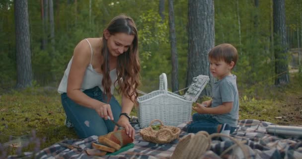 Młoda kobieta kroi chleb na pikniku, mały chłopiec siedzi na kocu, rodzina odpoczywa w lesie latem — Wideo stockowe