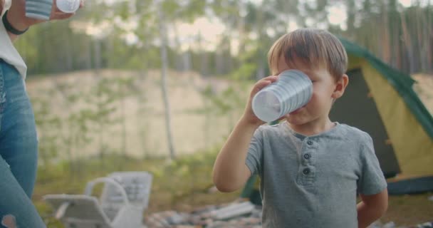Niño pequeño está bebiendo agua de vidrio en campamento de tienda en el bosque en verano, familia descansando en la naturaleza — Vídeos de Stock