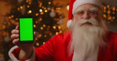 Noel Baba, Noel ağacının arkasındaki koltukta oturuyor ve elinde yeşil ekranlı bir cep telefonu olan çelenkler parmağıyla onu işaret ediyor..