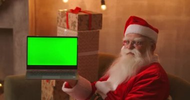Noel Baba, Noel süslemelerinde otururken elinde yeşil ekranlı bir laptop tutuyor..