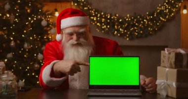 Noel ağacının arka planında, yaşlı bir Noel Baba dizüstü bilgisayar tutuyor ve bir kromatonla ekranı işaret ediyor. Yeşil ekranlı bir laptop. Noel Baba kameraya bakıyor..