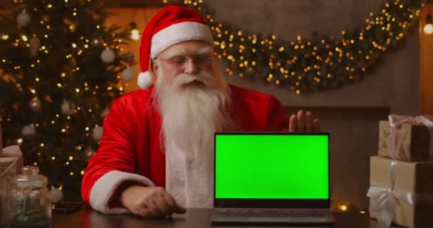 Santa sostiene un ordenador portátil con una pantalla verde mientras está sentado en las decoraciones de Navidad. — Vídeo de stock