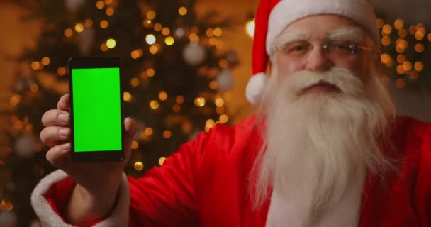 Santa Claus está sentado en el sofá en el fondo de un árbol de Navidad y guirnaldas sosteniendo un teléfono móvil con una pantalla verde apuntándolo con su dedo. — Vídeo de stock