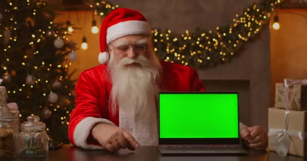 На заднем плане рождественской елки пожилой Санта-Клаус держит ноутбук и указывает на экран с хромакеем. Ноутбук с зеленым экраном. Санта смотрит в камеру. — стоковое видео