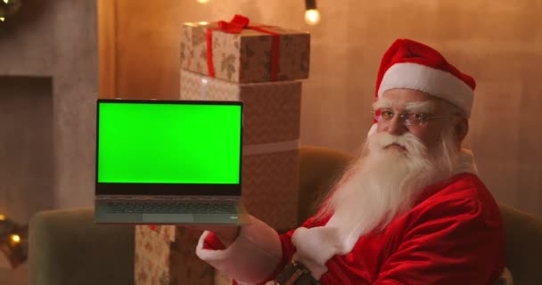 Di latar belakang pohon Natal, Santa Claus tua memegang laptop dan menunjuk ke layar dengan kromakey. Laptop dengan layar hijau. Santa melihat ke kamera. — Stok Video