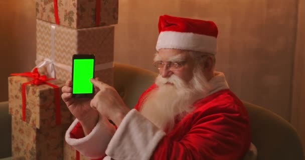 Santa δείχνει το δάχτυλό του στην πράσινη οθόνη του κινητού τηλεφώνου. Χριστουγεννιάτικη πώληση. Ηλικιωμένοι Άγιος Βασίλης δείχνει μια οθόνη με ένα chromakey — Αρχείο Βίντεο