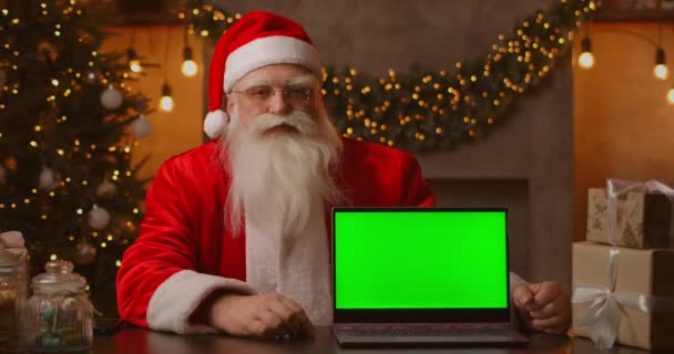 Santa drží notebook se zelenou obrazovkou, zatímco sedí v vánoční dekorace.