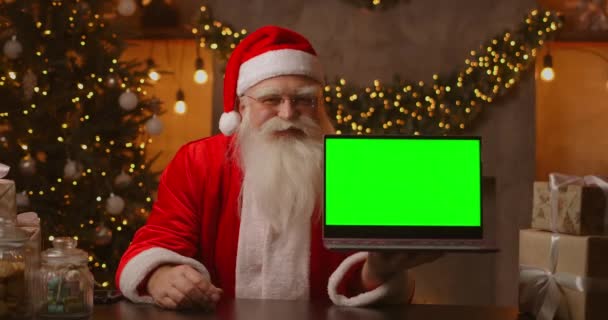 Op de achtergrond van de kerstboom houdt een bejaarde kerstman een laptop vast en wijst met een chromakey naar het scherm. Laptop met een groen scherm. Kerstman kijkt naar de camera. — Stockvideo