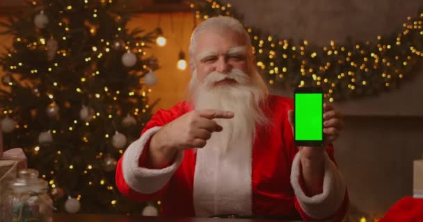 サンタクロースはクリスマスツリーの背景にソファの上に座っており、緑の画面が彼の指でそれを指している携帯電話を持っているガーランド. — ストック動画