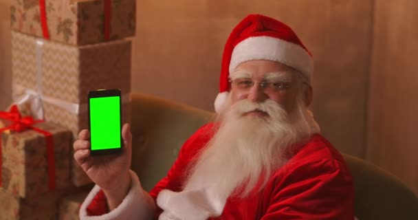 Święty Mikołaj siedzi na kanapie w tle choinki i girlandy trzymając telefon komórkowy z zielonym ekranem wskazując na nią palcem. — Wideo stockowe