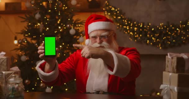 Санта показывает пальцем на зеленый экран мобильного телефона. Рождественская распродажа Пожилой Санта Клаус показывает экран с хромакеем — стоковое видео