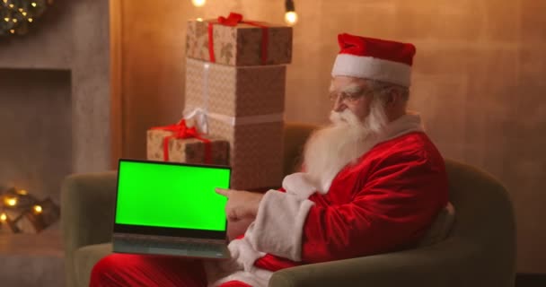 W tle choinki, starszy Święty Mikołaj trzyma laptopa i wskazuje na ekran chromakey. Laptop z zielonym ekranem. Mikołaj patrzy w kamerę.. — Wideo stockowe