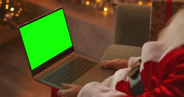 Santa Claus está hablando en una videollamada vía laptop. Utilice la pantalla verde para una videollamada. Santa Claus saluda con su mano mirando la pantalla verde del ordenador portátil — Vídeos de Stock