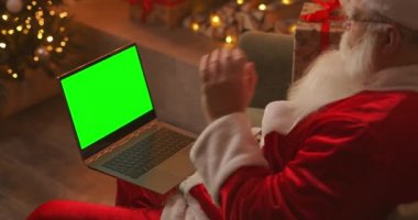 Noel Baba bilgisayarla video üzerinden konuşuyor. Görüntülü arama için yeşil ekranı kullan. Noel Baba yeşil dizüstü bilgisayara bakarak selamlıyor.