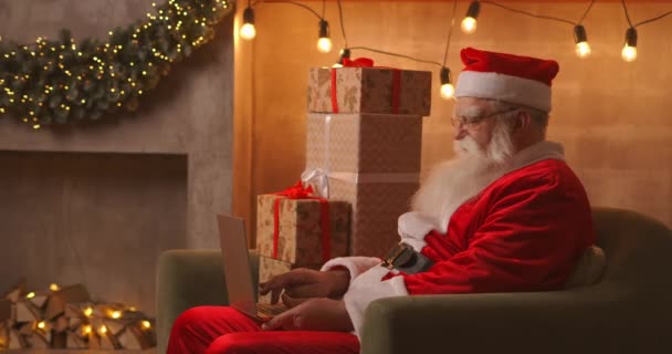 Santa Claus sedang mengerjakan laptop duduk di sofa dengan latar belakang Natal. Santa menjawab email anak-anak — Stok Video