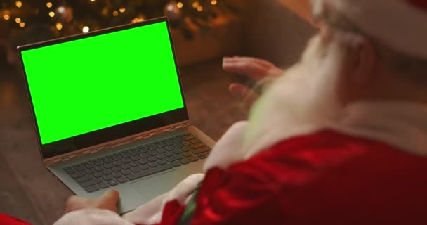 A Mikulás egy videóhíváson beszél laptopon keresztül. Használd a zöld képernyőt egy videohíváshoz. Mikulás üdvözöl a kezét nézi a zöld laptop képernyőn — Stock videók