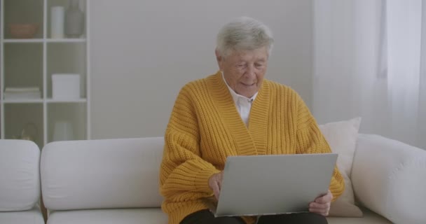 头发灰白的老年妇女在家里用笔记本电脑打视频电话。老年妇女和家人一起打了一个视频电话，面带微笑地挥手。COVID 19保持连接。与朋友在线聊天 — 图库视频影像