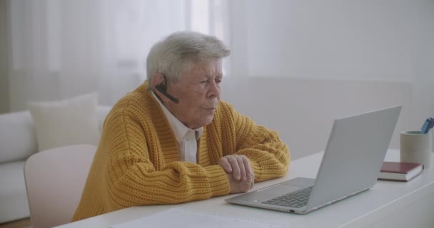 Seniorin mit Laptop telefoniert zu Hause mit Smartphone. Alte Frau mit einem Videoanruf auf dem Laptop lächelt und spricht fröhlich drinnen in einer gemütlichen Wohnung. Arztvideo mit älteren Patienten — Stockvideo