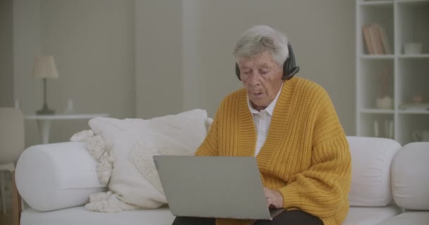 Senior kvinna med grått hår gör videosamtal med bärbar dator hemma. Äldre kvinna som har ett videosamtal med familj, leende och vinkande. COVID 19 Håll kontakten. Online chattar med vänner — Stockvideo