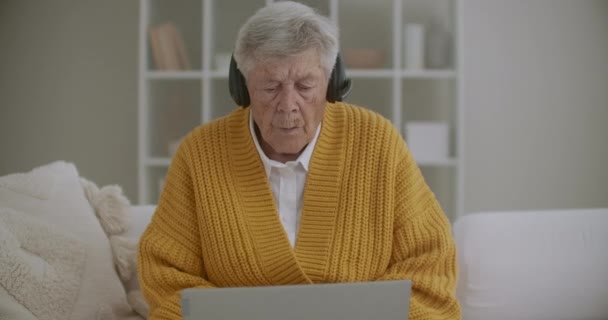 头发灰白的老年妇女在家里用笔记本电脑打视频电话。老年妇女和家人一起打了一个视频电话，面带微笑地挥手。COVID 19保持连接。与朋友在线聊天 — 图库视频影像
