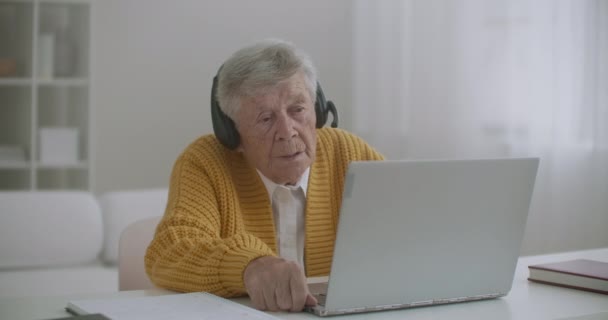 Seniorin mit Laptop telefoniert zu Hause mit Smartphone. Alte Frau mit einem Videoanruf auf dem Laptop lächelt und spricht fröhlich drinnen in einer gemütlichen Wohnung. Arztvideo mit älteren Patienten — Stockvideo