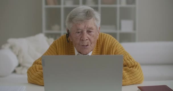 有笔记本电脑的老年妇女在家里用智能手机打电话。老妇人笑着在笔记本电脑上打了一个视频电话，在一个舒适的公寓里愉快地聊天。医生视频呼叫老年患者 — 图库视频影像