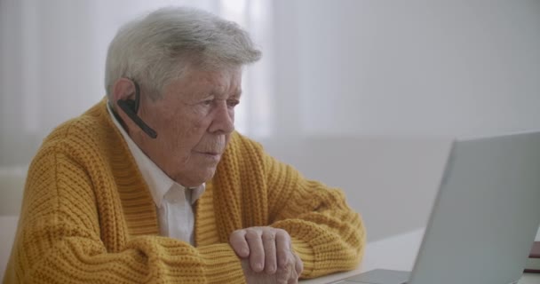自宅でスマートフォンを呼び出すノートパソコンのコンピュータを持つシニア女性。ラップトップでビデオ通話をしている老婦人は笑顔で、居心地の良いアパートで屋内で楽しく話しています。高齢患者と呼ばれる医師のビデオ — ストック動画