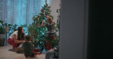 Bir anne ve iki çocuk evlerinin oturma odasında bir Noel ağacını oyuncaklarla süslüyor. Noel süslemeleri ve tatil hazırlıkları.