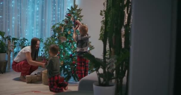 Eine junge Mutter und zwei Jungen im Pyjama schmücken an Silvester den Weihnachtsbaum mit Spielzeug. Vorbereitung auf das neue Jahr und Weihnachten. Das Haus schmücken — Stockvideo