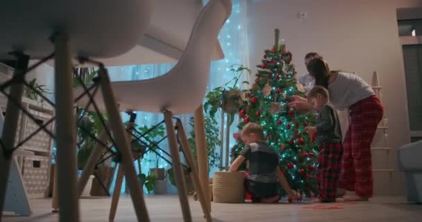 Sebuah keluarga dengan dua anak menghias pohon Natal pada malam Natal. Ayah dan ibu dengan anak-anak mempersiapkan untuk tahun baru dan rumah dekorasi Natal — Stok Video