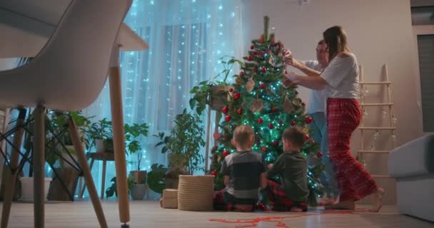 新年の冬のお祝いのコンセプトのための幸せな家族の準備。若い30代のカップル彼の就学前の愛らしい2人の息子装飾クリスマスツリー作成お祝いムード雰囲気で現代居心地の良い — ストック動画