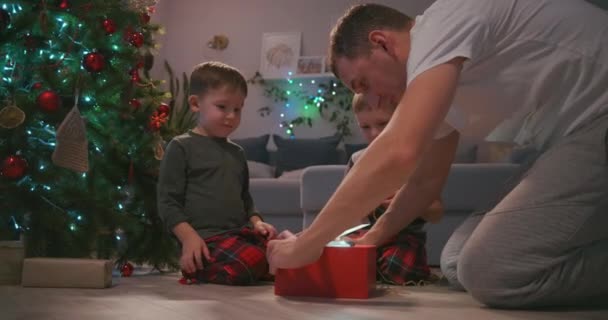 Père avec deux enfants ouvre un cadeau de Noël sous le sapin et se réjouit. Un cadeau pour mon père. Un rayon de lumière du cadeau brille sur le visage. Réjouissez-vous avec les enfants cadeau du Nouvel An. — Video