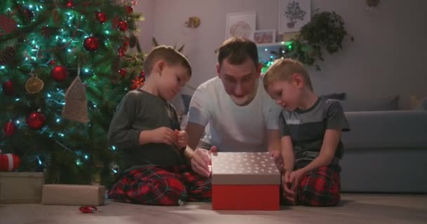 Vater mit zwei Kindern öffnet ein Weihnachtsgeschenk unter dem Baum und freut sich. Ein Geschenk für meinen Vater. Ein Lichtstrahl des Geschenks leuchtet ins Gesicht. Freuen Sie sich mit den Kindern über das Neujahrsgeschenk. — Stockvideo
