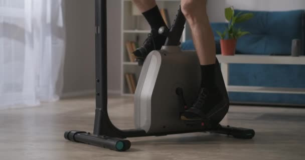 Evde yalnızlık sırasında spor, erkek bacakları oturma odasında sabit bisiklet pedalları çeviriyor, yakın görüş, evde antrenman yapıyor. — Stok video
