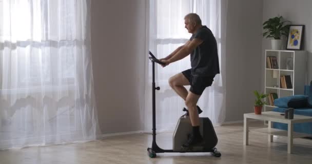 Σύγχρονο σταθερό ποδήλατο στο σπίτι, ενήλικας άνθρωπος είναι η κατάρτιση σχετικά με τον εξοπλισμό γυμναστικής, απώλεια βάρους — Αρχείο Βίντεο