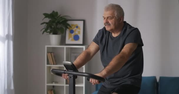 Man van middelbare leeftijd traint met stationaire fiets in huis, cardiotraining, het houden van lichaamsbeweging — Stockvideo