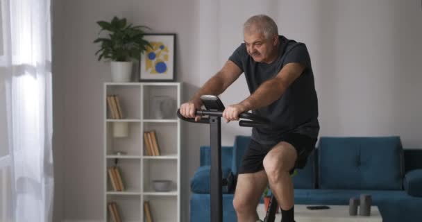 Medelålders man tränar med motionscykel hemma, medium skott av gråhårig person med mustasch — Stockvideo