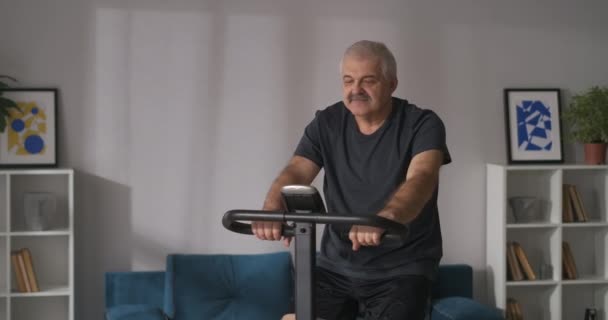 Portret van middelbare leeftijd man op stationaire fiets in appartement, training thuis, het houden van een goede lichamelijke conditie — Stockvideo