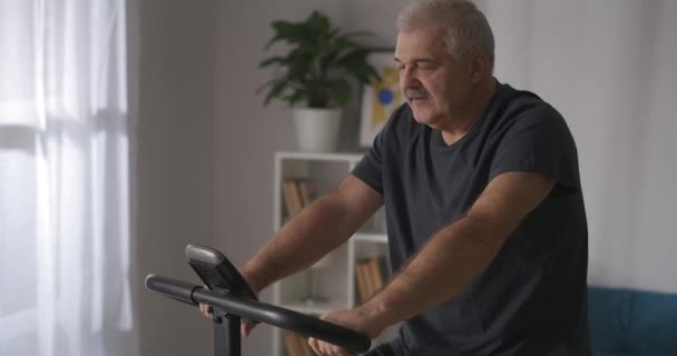 Portrait of middle-aged man, training with stationary bike at home, caring about health, keeping fit — Stock Video