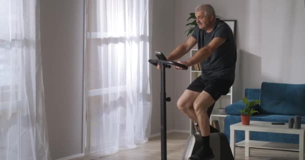 Σπορ μεσήλικας άνδρας προπονείται με σταθερό ποδήλατο στο σπίτι, μέτρια βολή στο σαλόνι — Αρχείο Βίντεο
