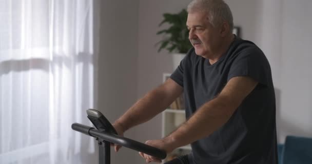 Homme d'âge moyen s'entraîne seul à la maison, en utilisant un vélo stationnaire moderne pour l'entraînement, en gardant la forme dans la vieillesse — Video