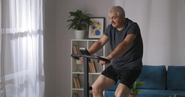 Középkorú férfi forog pedálok modern álló kerékpár otthon és megtekintése videó a képernyőn, kardió képzés — Stock videók